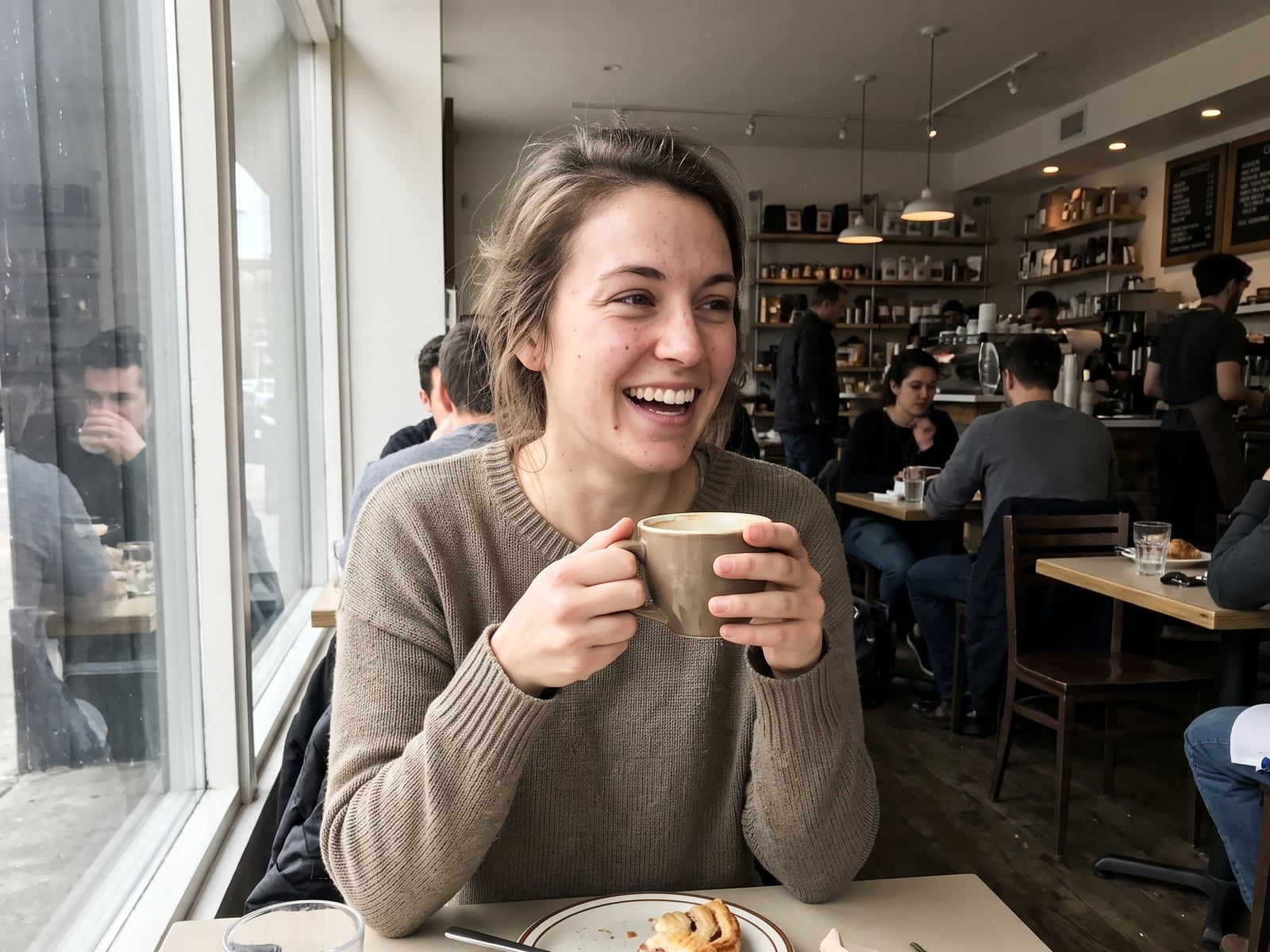 A young woman in a sweater holding a coffee and smiling across a table by a cafe window, other customers behind her